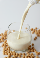ガラスの丸いコップに注がれている豆乳。周りには大豆が散らばっている。