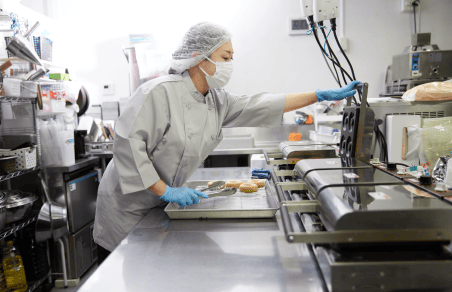 工場でドーナツを作っている様子。ドーナツを焼く専用の機械を使って、スタッフがドーナツを一つ一つ焼いている。スタッフはマスクやキャップ、手袋などを身につけ、衛生管理も徹底している。