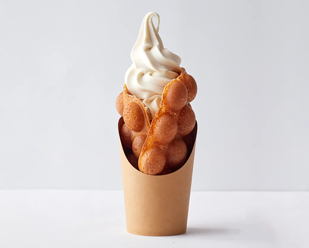 ワッフルソフト。きつね色に焼かれたバブルワッフルが軽く丸められており、その間に白いソフトクリームが高く巻かれている。