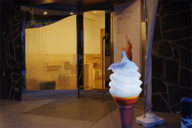 Soy storiesのお店の外の様子。ガラス戸の前にワッフルソフトの写真が印刷された幕が飾られており、その手前にはソフトクリームのライトが闇夜に輝いている。